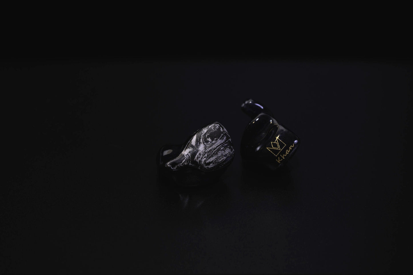 A pair of black custom in-ear monitors with a distinctive design, featuring a black and silver color scheme and a metallic finish. Right in-ear monitor is flipped to show a gold Noble logo and cursive "Khan".